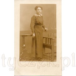Dystyngowana dama stojąca przy bardzo stylowych meblach: fotelu Art Deco oraz stoliku ludwikowskim